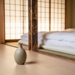 Japanese-inspired minimalist living room with a low platform bed, shoji-style screen filtering light, and a single ceramic vase on a light wood floor.